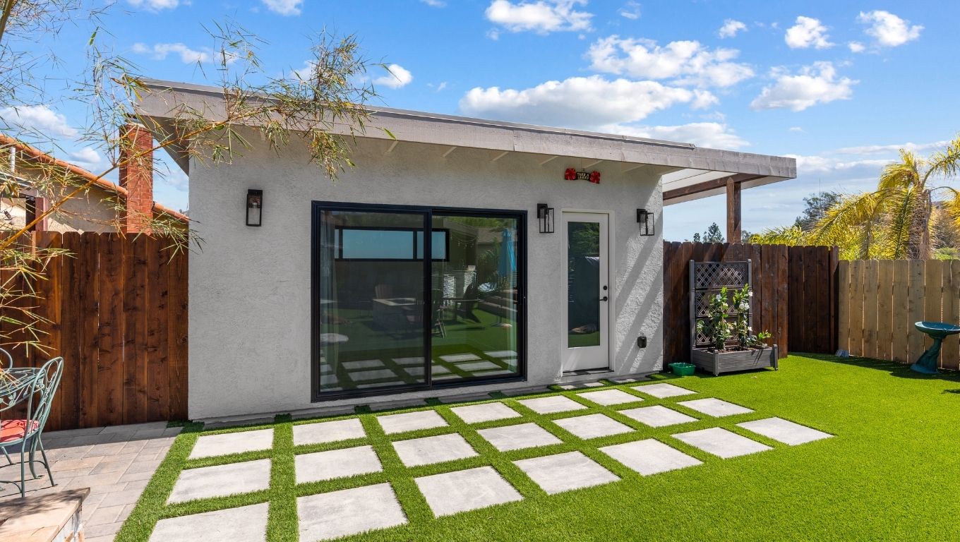 Modern backyard ADU exterior with white stucco, black-framed sliding doors, string lighting, and turf and paver yard