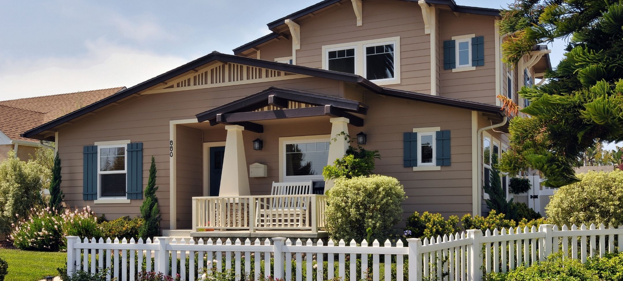 Craftsman-style custom home with tan siding, blue shutters, covered porch, and white picket fence