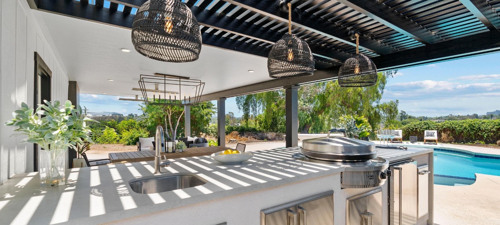 Covered outdoor kitchen and pergola with pendant lighting overlooking the backyard pool