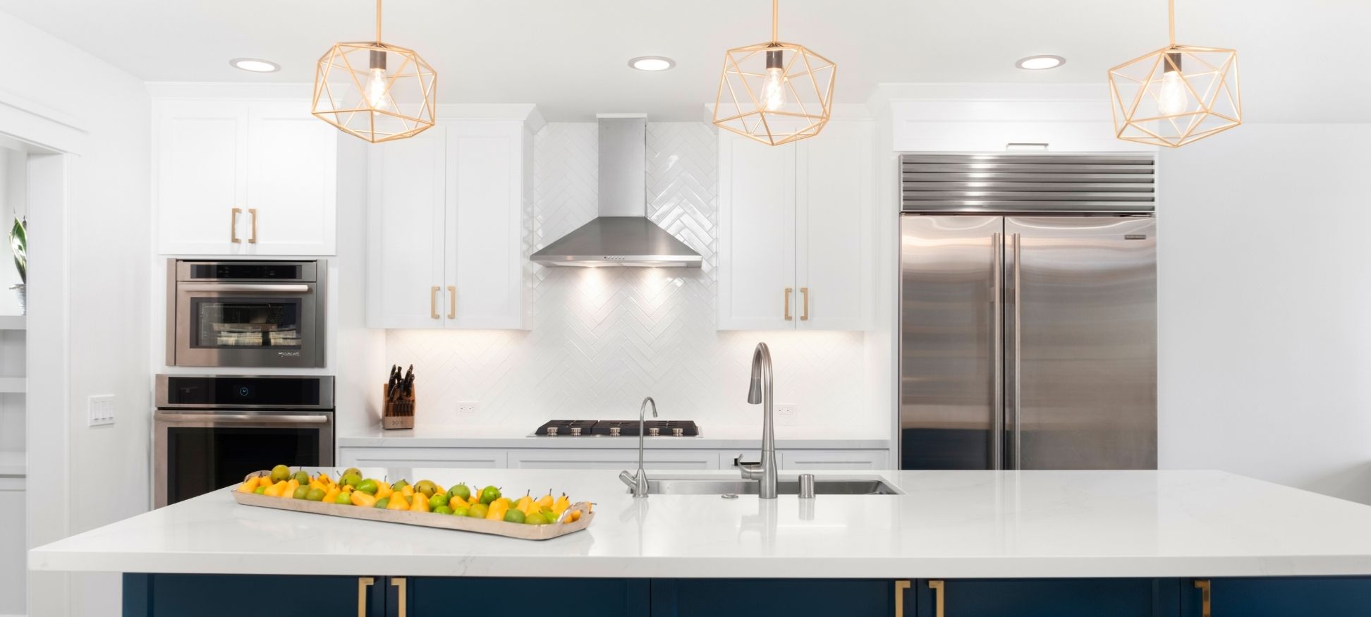 White kitchen remodel with gold geometric pendant lighting, navy blue island, and herringbone tile backsplash