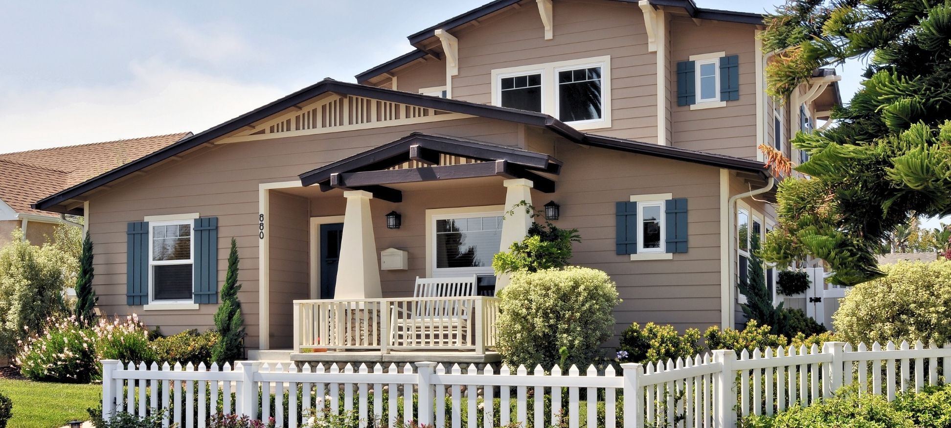 Two-story Craftsman-style home exterior with tan siding, blue shutters, and covered front porch with white picket fence