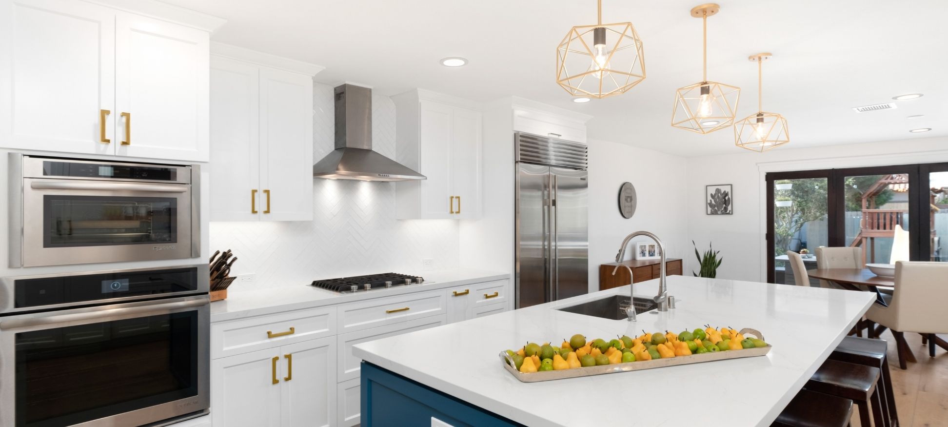 White kitchen remodel with herringbone tile backsplash, geometric brass pendant lighting, and navy blue island