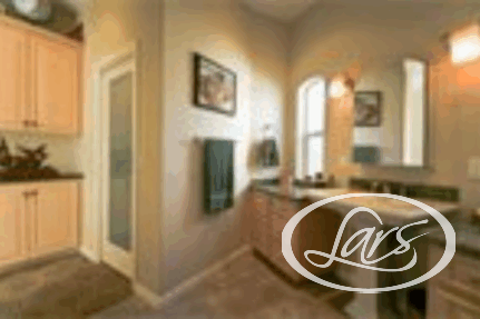 A modern bathroom featuring a pedestal sink with a large mirror above it, a countertop with storage cabinets, a framed window, and a towel hung on the wall. The room has beige walls and bright overhead lighting. A part of the adjacent room is also visible.
