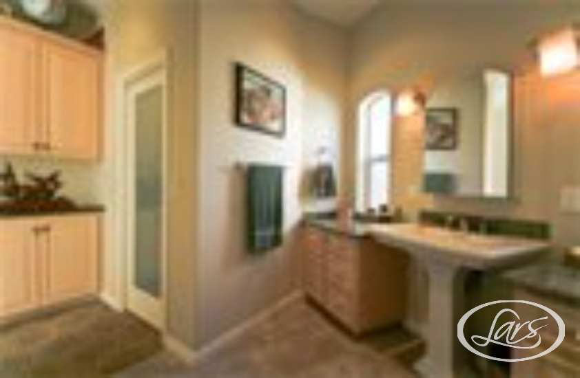 A modern bathroom featuring a pedestal sink with a large mirror above it, a countertop with storage cabinets, a framed window, and a towel hung on the wall. The room has beige walls and bright overhead lighting. A part of the adjacent room is also visible.