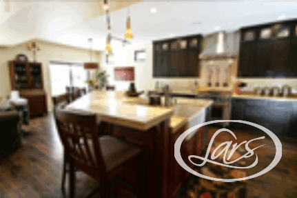 A modern kitchen with dark wooden cabinets, stainless steel appliances, and a large island with a light countertop. The kitchen island has two wooden bar stools and pendant lights hanging above. The room is well-lit by natural light from a large window.