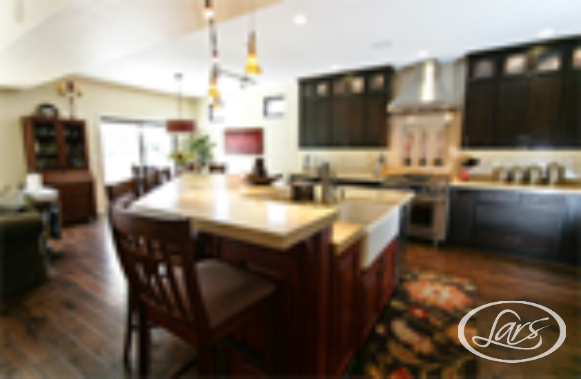A modern kitchen with dark wooden cabinets, stainless steel appliances, and a large island with a light countertop. The kitchen island has two wooden bar stools and pendant lights hanging above. The room is well-lit by natural light from a large window.