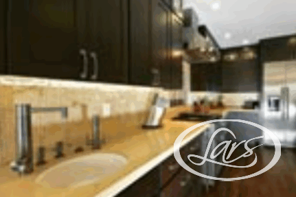 Modern kitchen with dark wood cabinets, light countertops, and stainless steel appliances. There is a built-in oven range with a vent hood and a refrigerator in the background. The kitchen is well-lit with under-cabinet lighting and recessed ceiling lights.