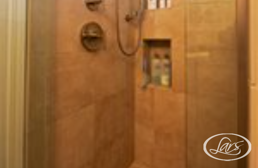 A modern shower area with beige tile walls and flooring. The shower features a wall-mounted shower head, a handheld spray, and built-in shelves with various toiletries. There is a glass door partially visible on the left side.