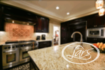 A modern kitchen with dark wooden cabinets, stainless steel appliances, and a large granite island countertop. The island features a built-in sink and a bowl of fresh fruit. The kitchen is illuminated by recessed lighting and pendant lights.