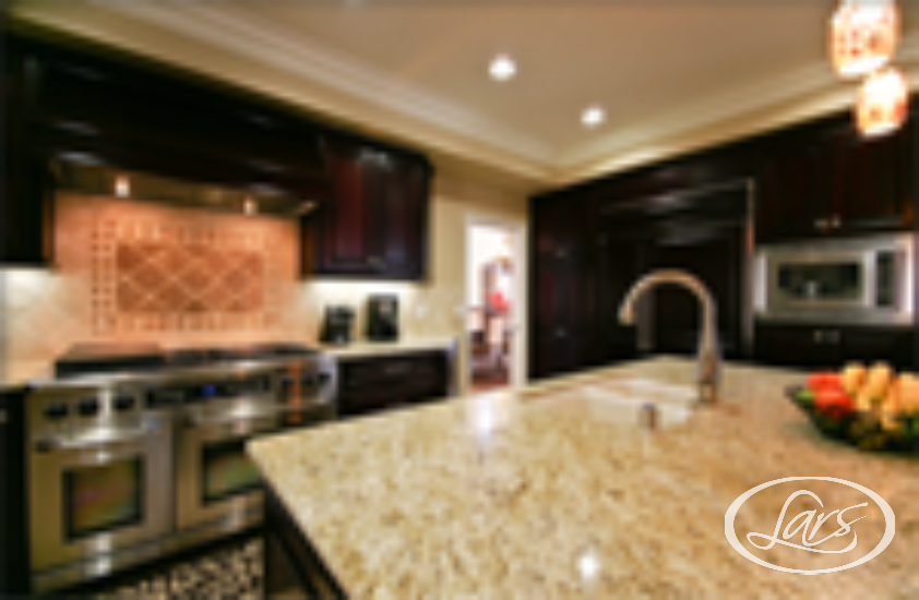 A modern kitchen with dark wooden cabinets, stainless steel appliances, and a large granite island countertop. The island features a built-in sink and a bowl of fresh fruit. The kitchen is illuminated by recessed lighting and pendant lights.