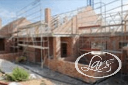A partially constructed brick house with scaffolding on the exterior. The roof framework is visible, and building materials are scattered around the site. The sky is clear, indicating daytime.
