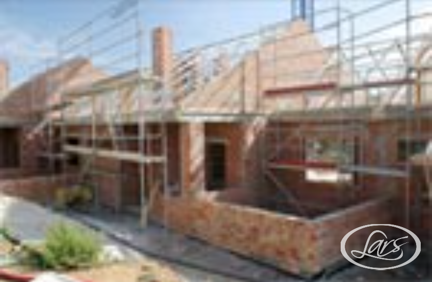 A partially constructed brick house with scaffolding on the exterior. The roof framework is visible, and building materials are scattered around the site. The sky is clear, indicating daytime.