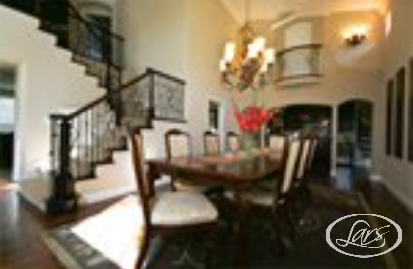 An elegant dining room featuring a long wooden table with six chairs, a chandelier overhead, and a staircase with ornate railings. The room has high ceilings, a large arched doorway, and ambient lighting, creating a sophisticated atmosphere.