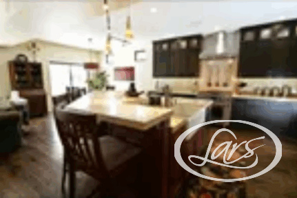 A modern kitchen with dark wooden cabinets and stainless steel appliances. There is a large island in the center with bar stools, a rug beneath it, and pendant lights hanging above. An adjacent dining area and a living space are partially visible in the background.