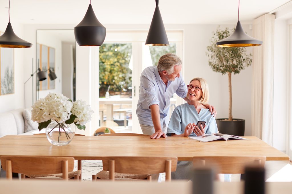 Senior Couple Relaxing With Magazine At Home Looking At Mobile Phone Sitting At Dining Room Table