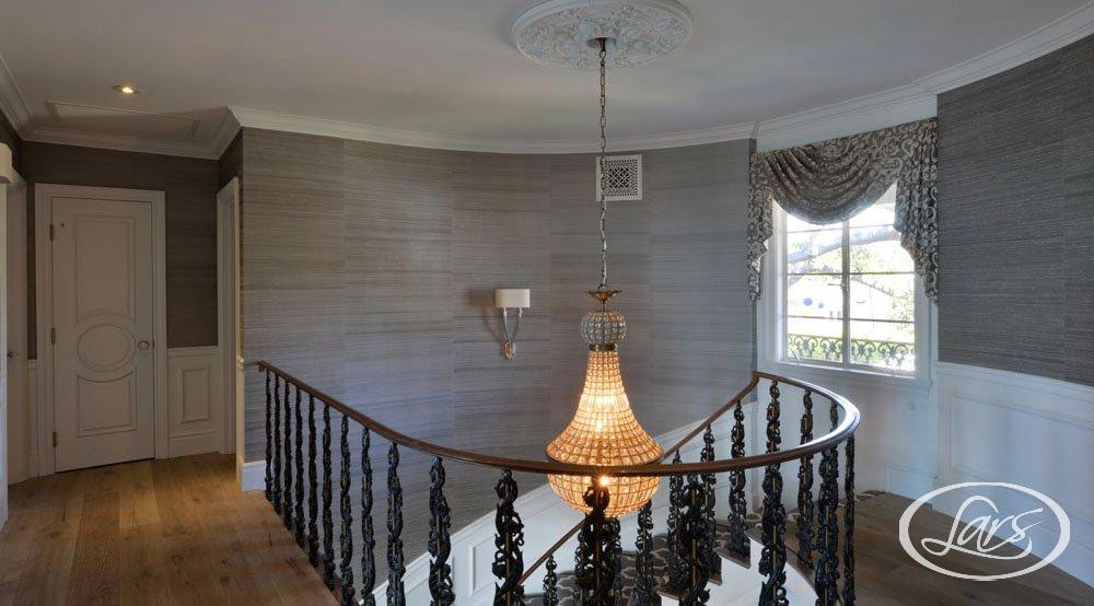A spiral staircase with ornate black railings and a large chandelier hangs from the ceiling. The walls are covered in textured gray wallpaper, and there is a single sconce on the wall. A window dressed with drapes allows natural light to brighten the space.