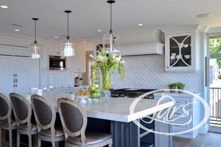 A modern kitchen with white cabinetry, a large island with a light marble countertop, and four upholstered barstools. Pendant lights hang above the island. Appliances include a stove, oven, and microwave. A vase of lilies and a bowl of green apples sit on the island.