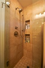 A modern shower area with beige tile walls and flooring. The shower features a wall-mounted shower head, a handheld spray, and built-in shelves with various toiletries. There is a glass door partially visible on the left side.