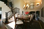An elegant dining room featuring a long wooden table with six chairs, a chandelier overhead, and a staircase with ornate railings. The room has high ceilings, a large arched doorway, and ambient lighting, creating a sophisticated atmosphere.