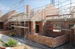 A partially constructed brick house with scaffolding on the exterior. The roof framework is visible, and building materials are scattered around the site. The sky is clear, indicating daytime.