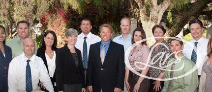 A diverse group of 13 people in formal attire stand together outdoors. They are smiling and positioned in a line, with trees and greenery in the background, suggesting a park or garden setting.