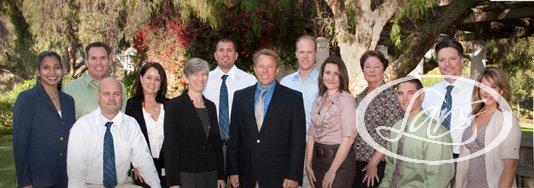 A diverse group of 13 people in formal attire stand together outdoors. They are smiling and positioned in a line, with trees and greenery in the background, suggesting a park or garden setting.