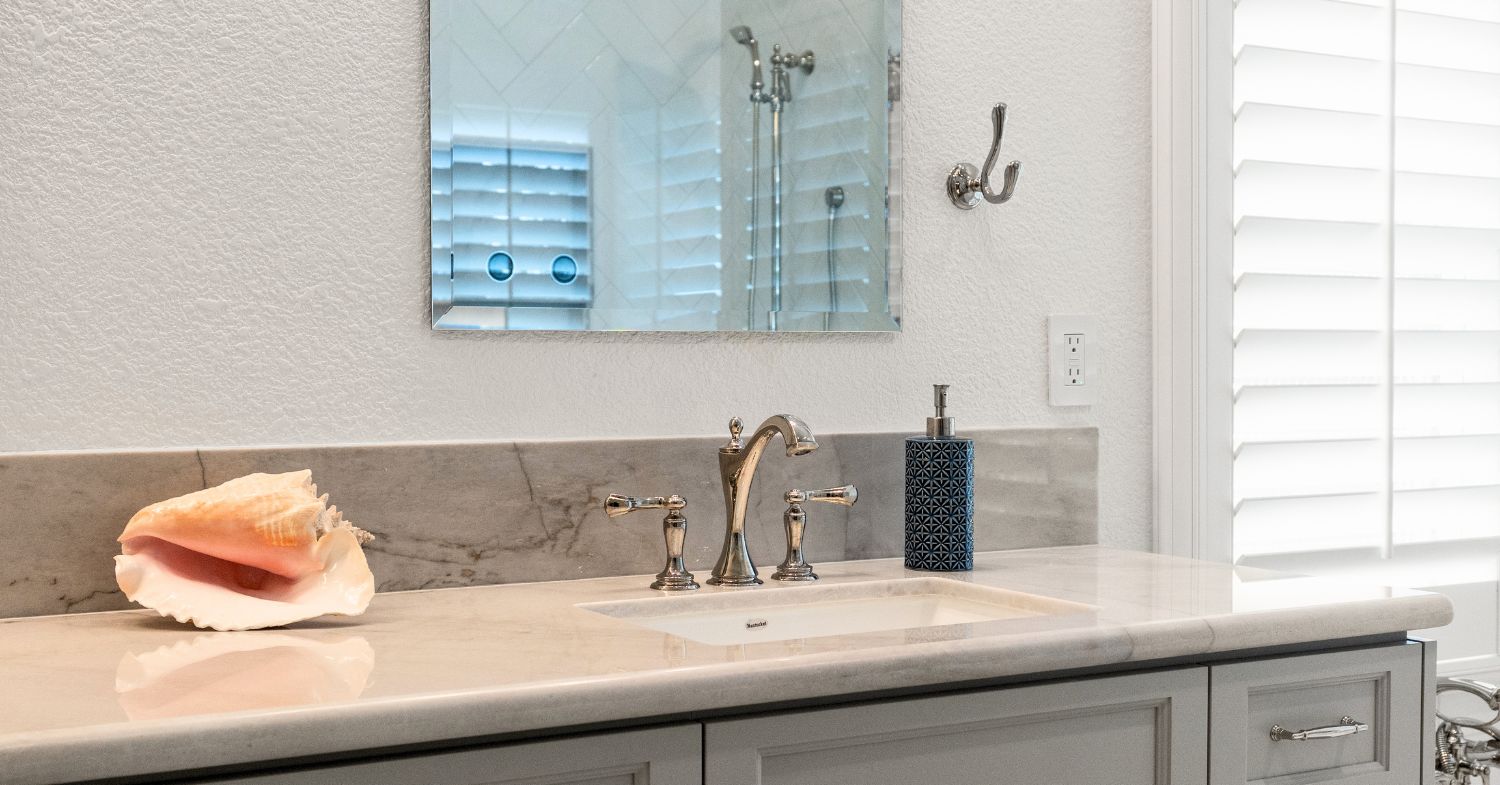 Carmel Valley bathroom remodel detail — marble countertop with chrome widespread faucet, gray shaker vanity, beveled mirror, and plantation shutters in a Carmel Valley home remodeled by Lars Remodeling & Design