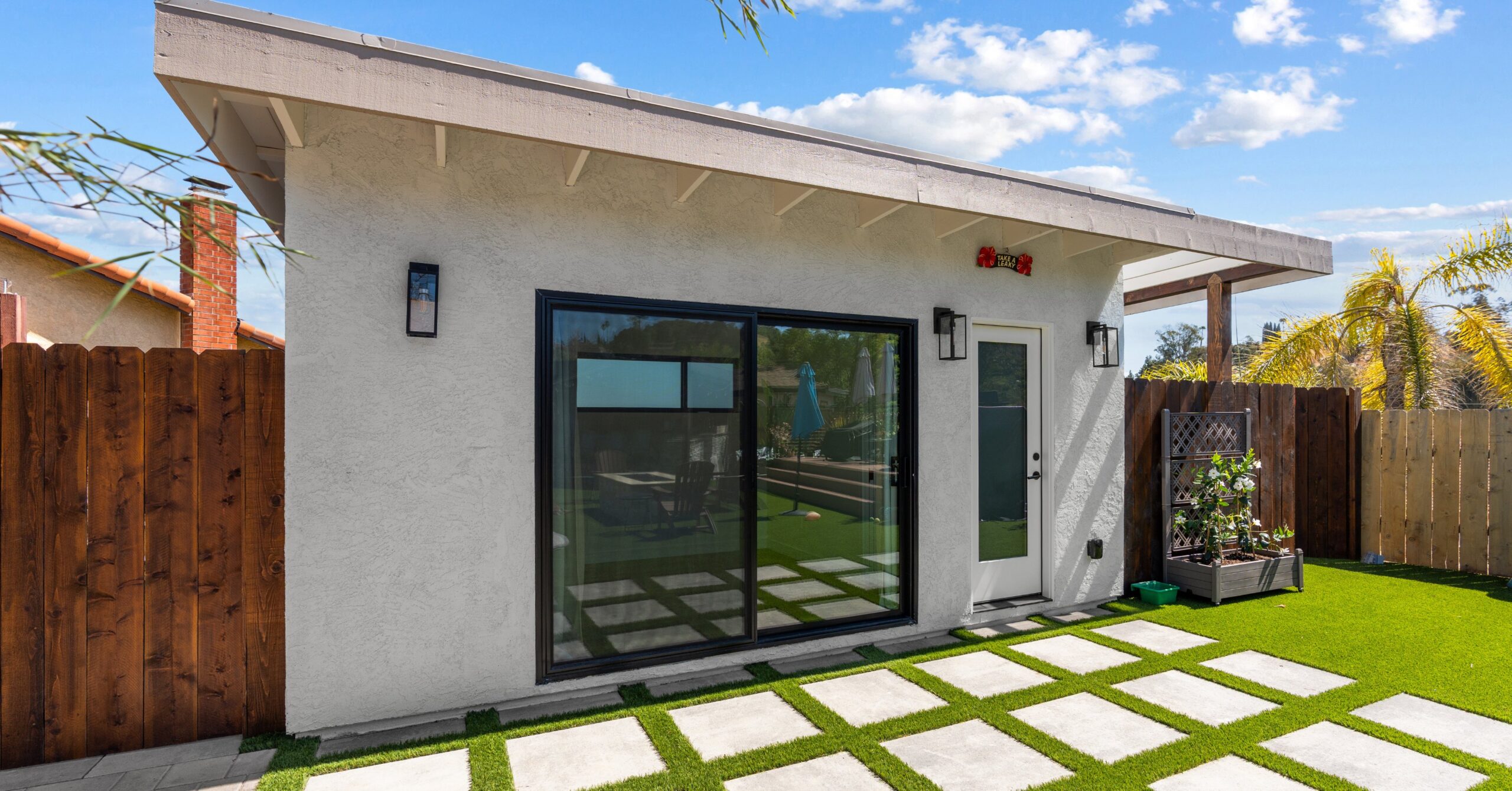 Exterior of a custom backyard accessory structure with sliding glass doors, modern lighting, paver pathways, and artificial turf in Spring Valley, California.
