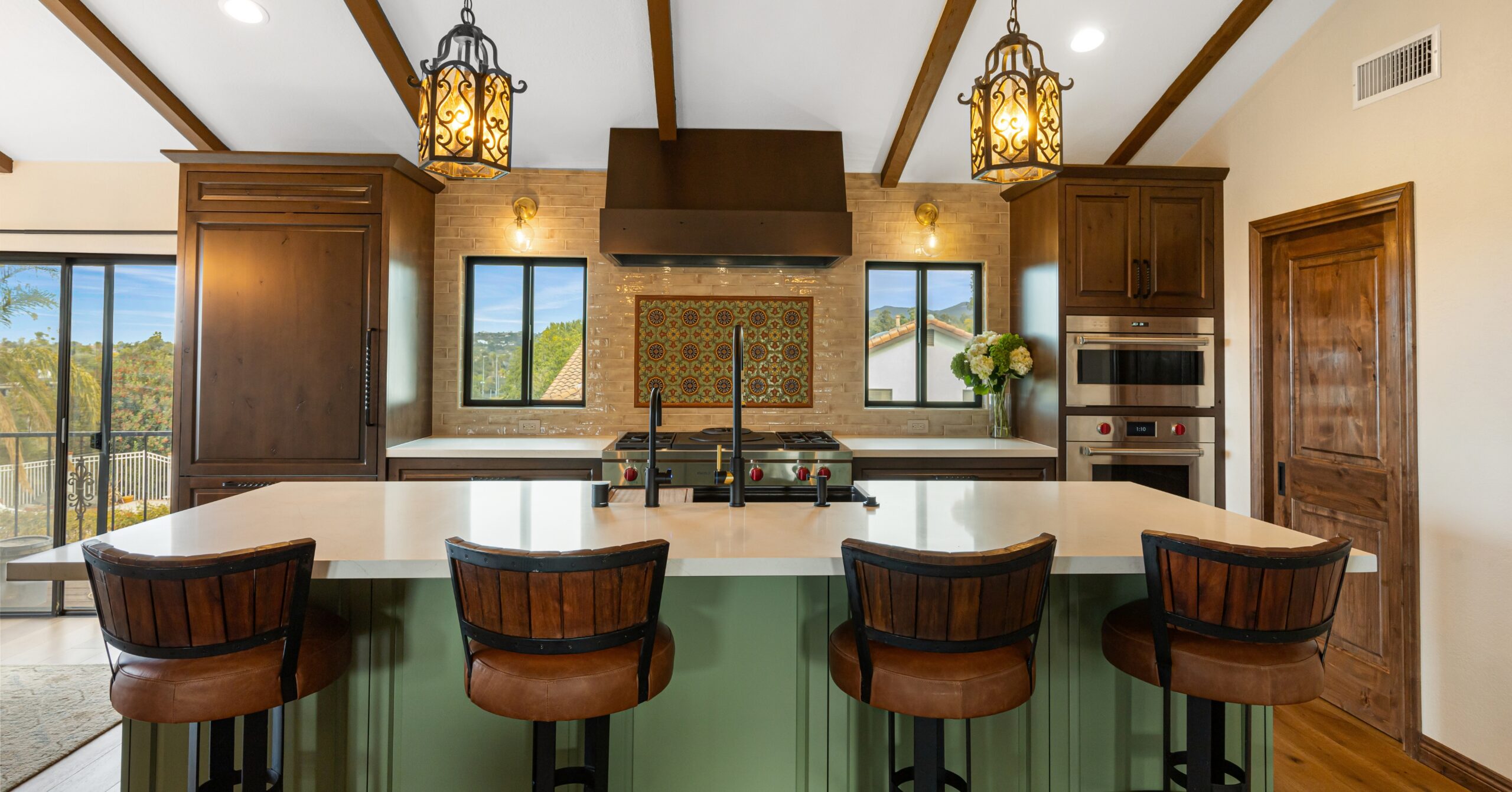 Mission-Spanish kitchen remodel in Vista CA featuring a large sage green island, warm wood cabinetry, vaulted ceilings with exposed beams, decorative lantern lighting, and custom tile backsplash.