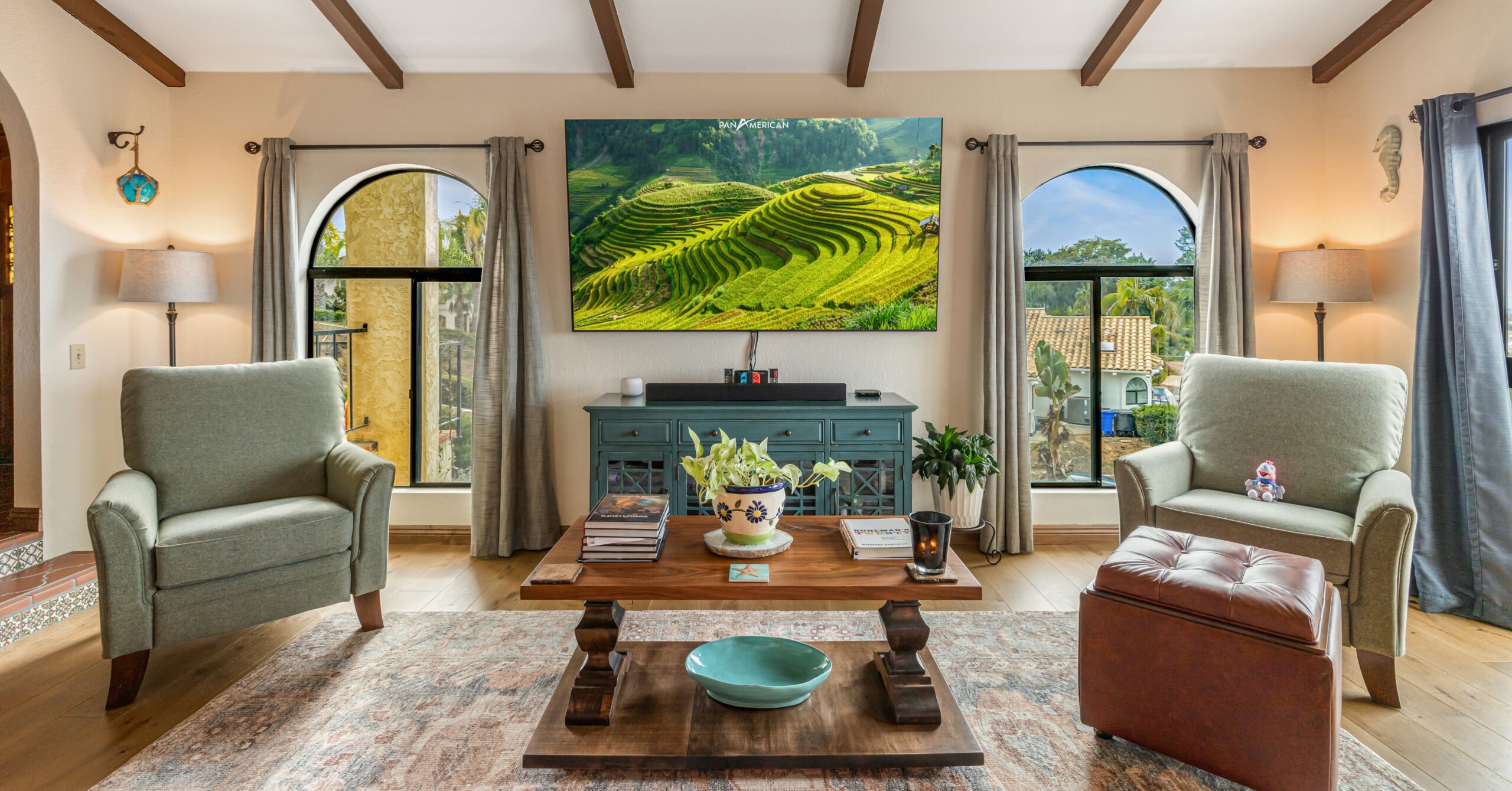 Connected living room adjacent to a remodeled kitchen featuring exposed ceiling beams, arched windows, warm wood flooring, and Mission-style design elements.