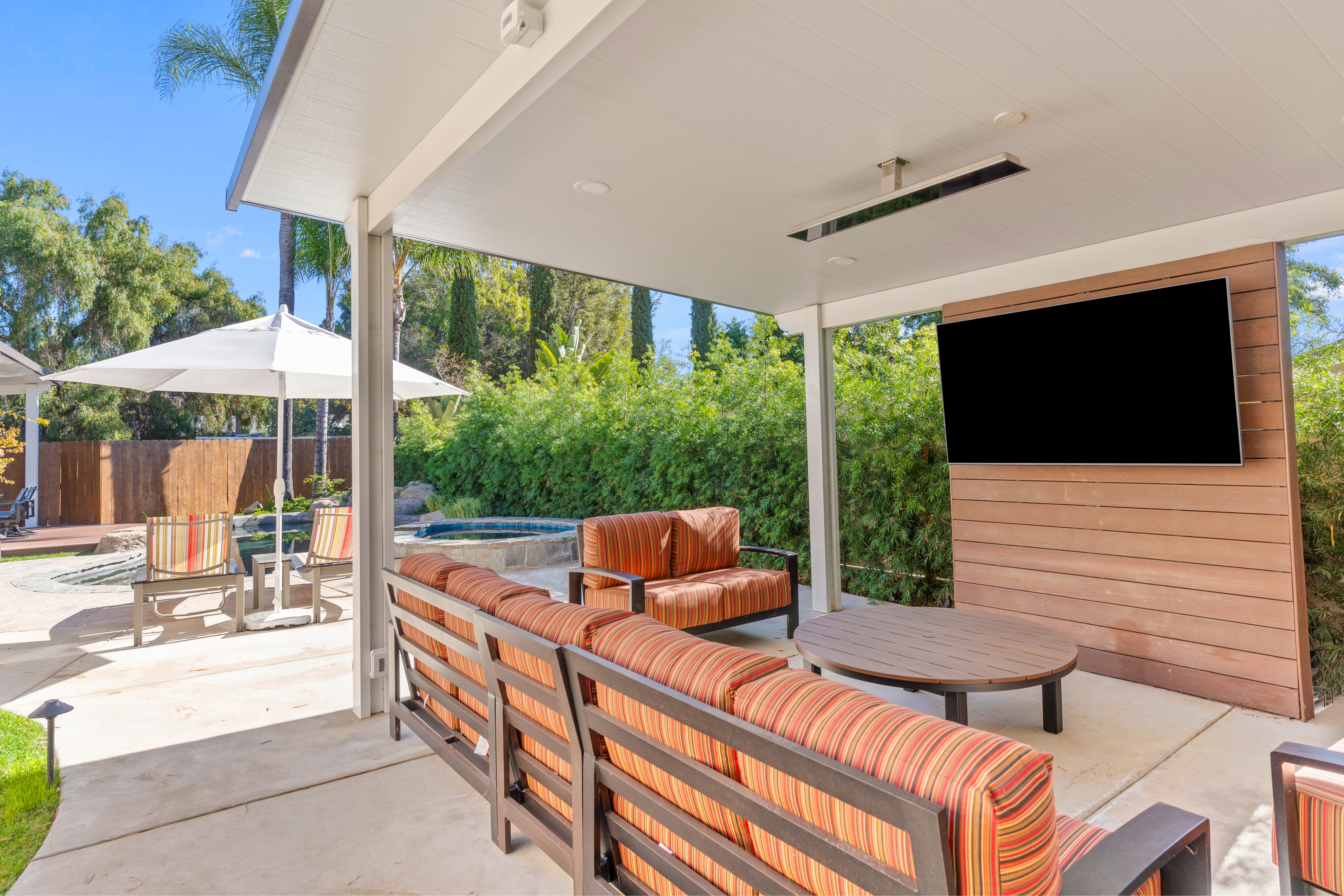 Covered poolside lounge with striped seating, mounted TV, wood-look accent wall and overhead heaters.