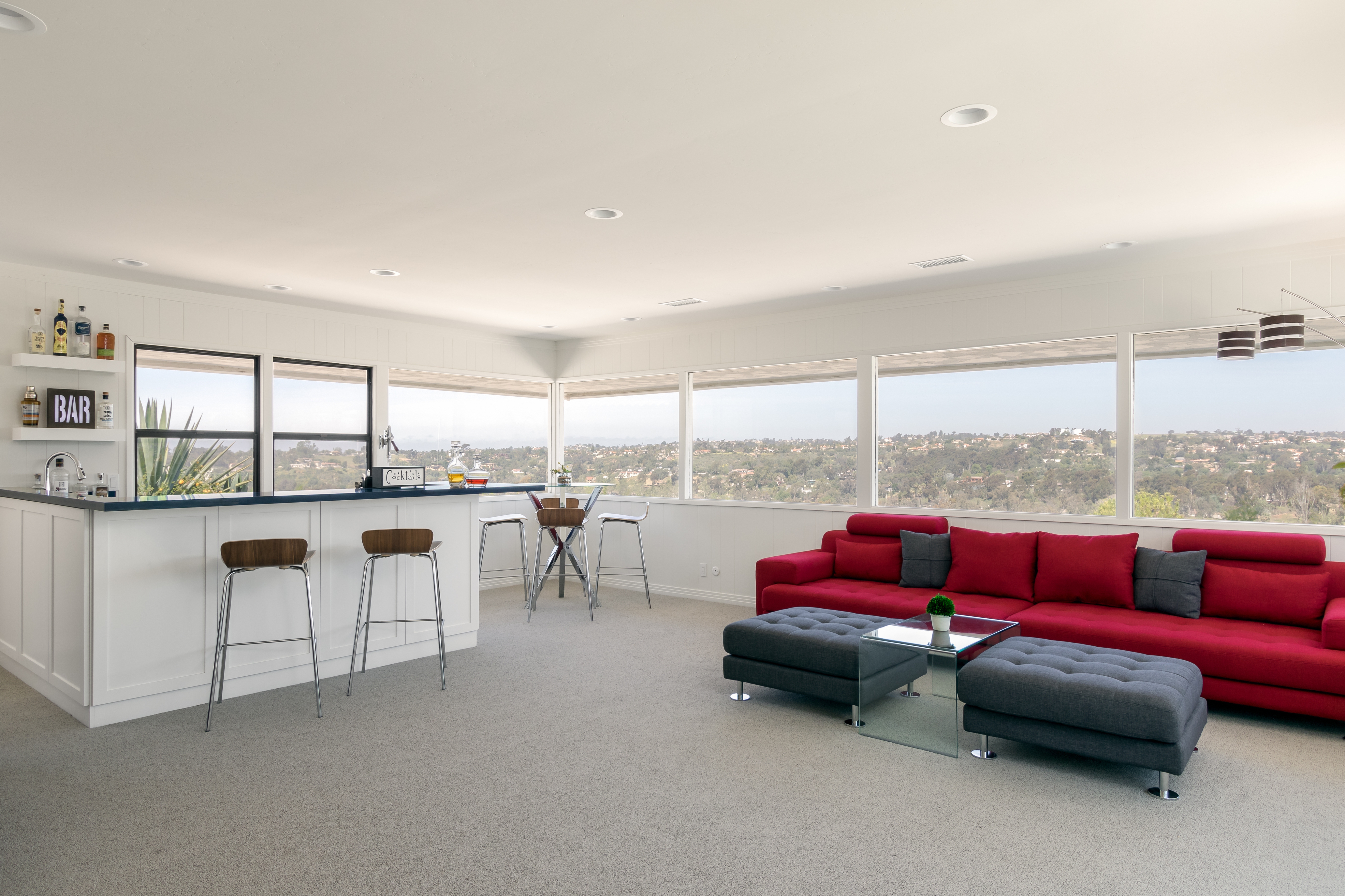 Open lounge with wet bar, panoramic windows, red sectional and gray ottomans.