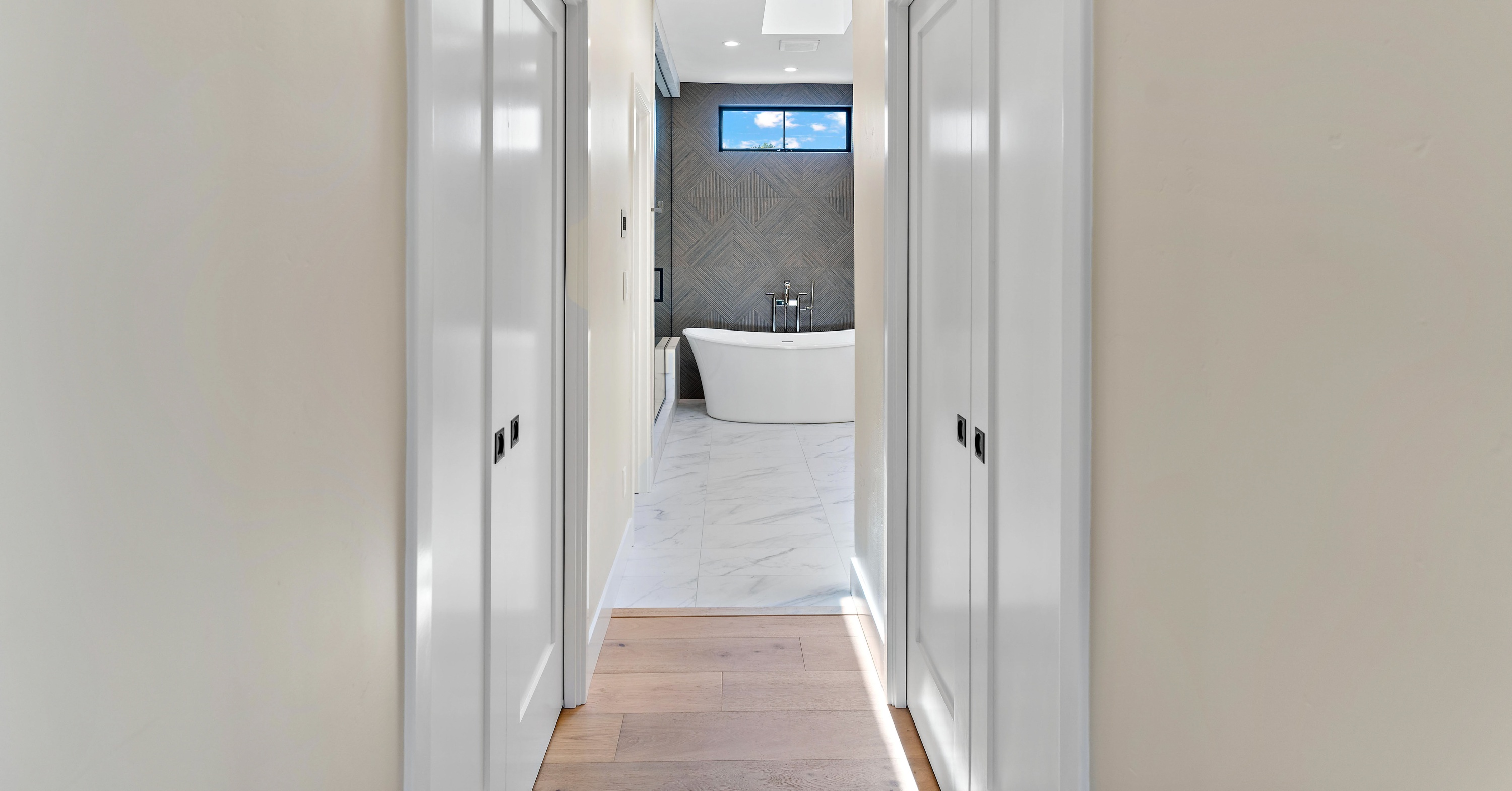 Walk-through closet connecting a primary bedroom to a luxury en suite bathroom with wood flooring, built-in storage, and a freestanding soaking tub.