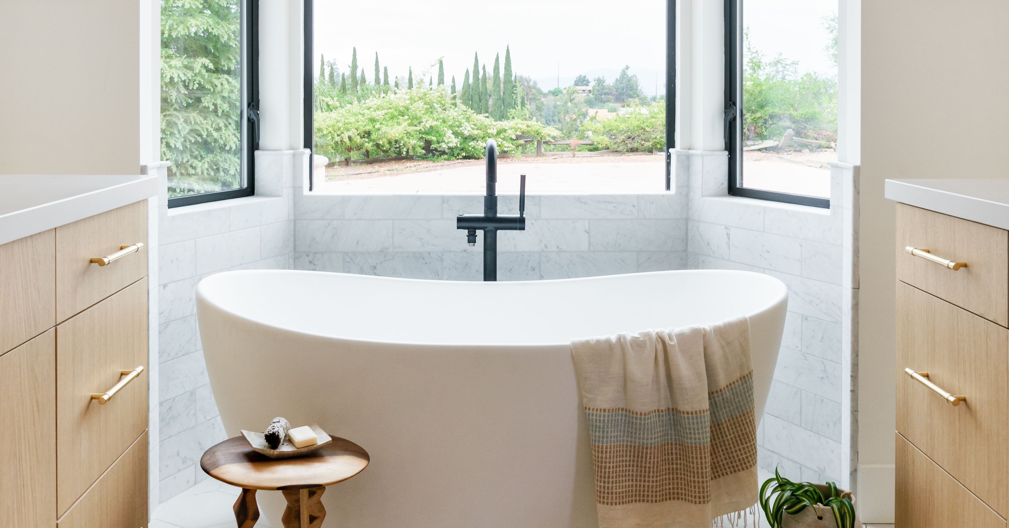 Freestanding soaking tub beneath bay windows with marble tile, light wood vanities, and a matte black faucet.
