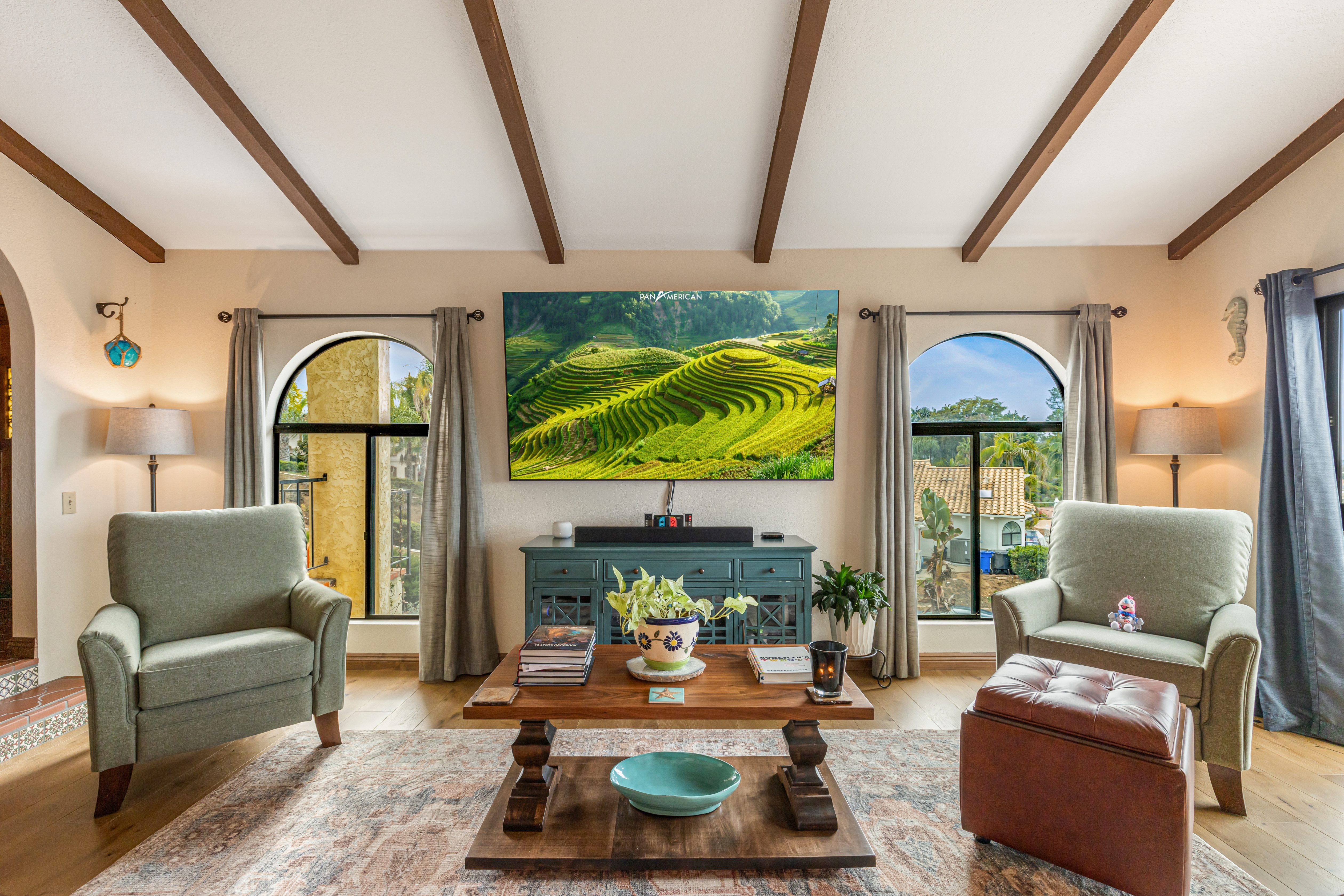 Living room with vaulted exposed-beam ceiling, arched windows, wood flooring and centered media wall.