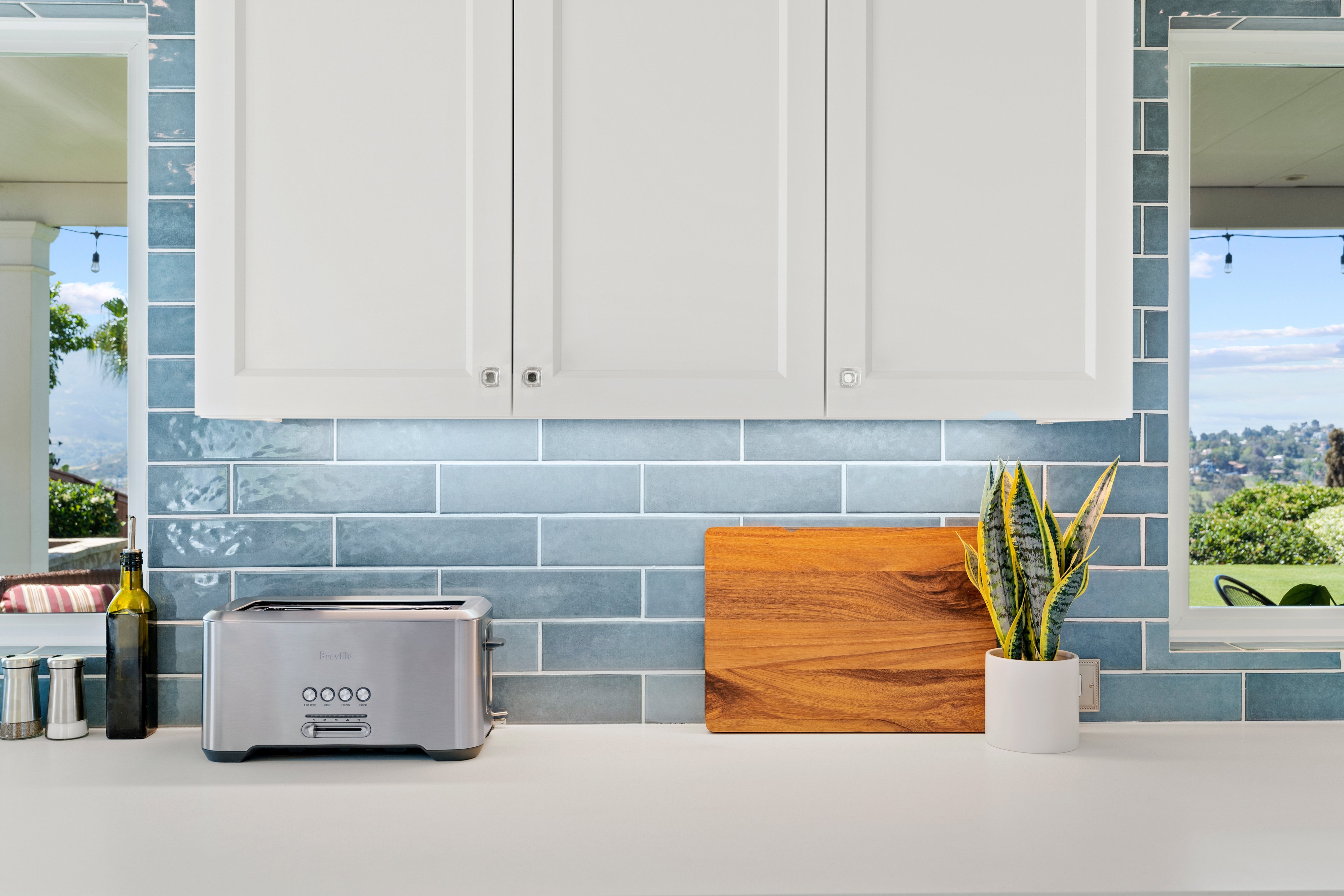 White upper kitchen cabinets above blue subway tile, a light countertop and two view windows.
