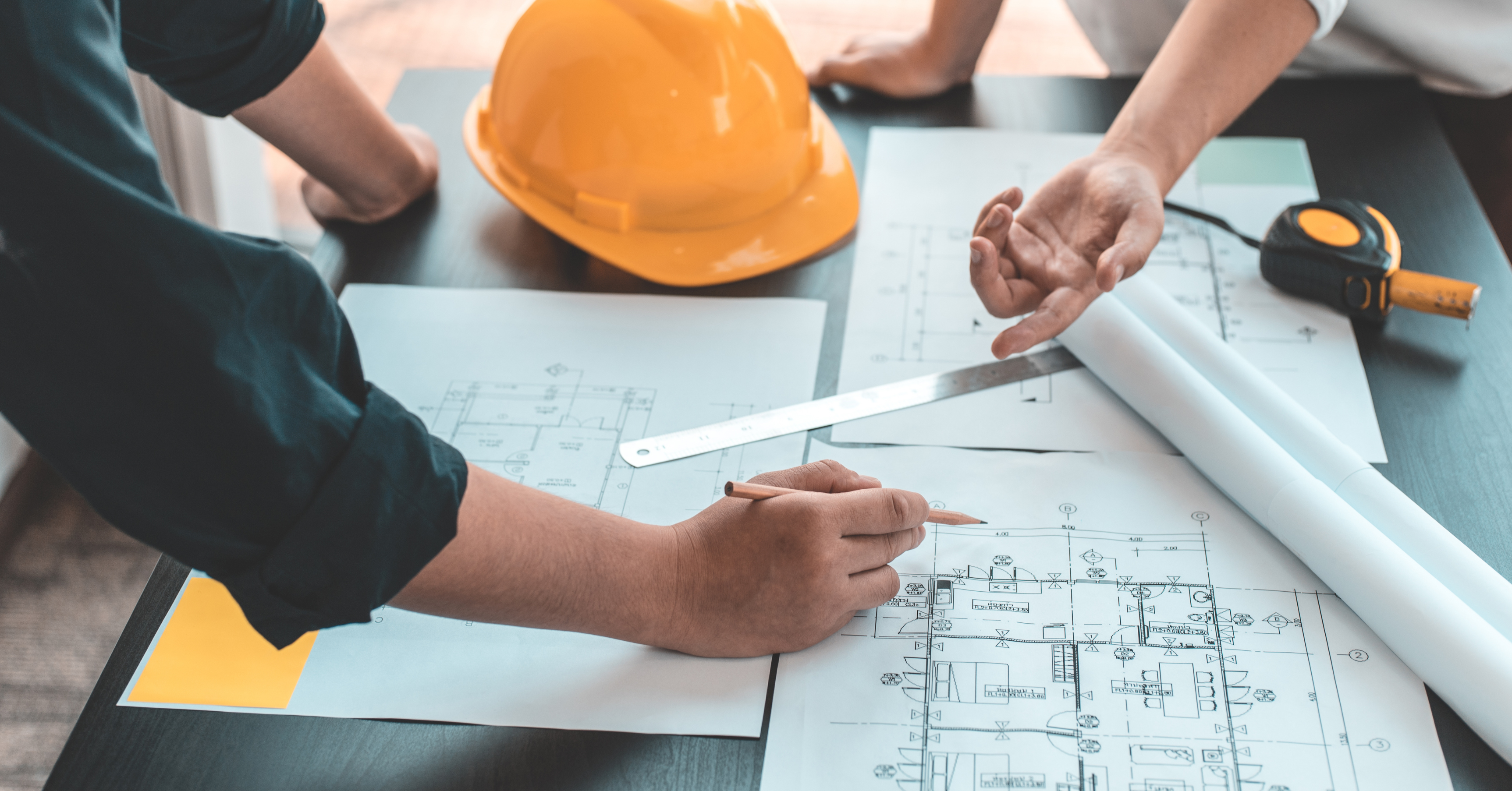 Homeowners reviewing architectural plans with a contractor, construction drawings, hard hat, and measuring tape during the renovation planning process.