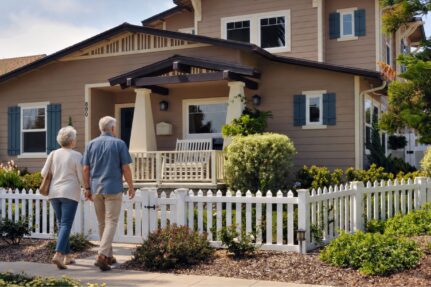 Older couple walking toward a well-maintained home with landscaped yard and front porch, representing independent living and aging in place.