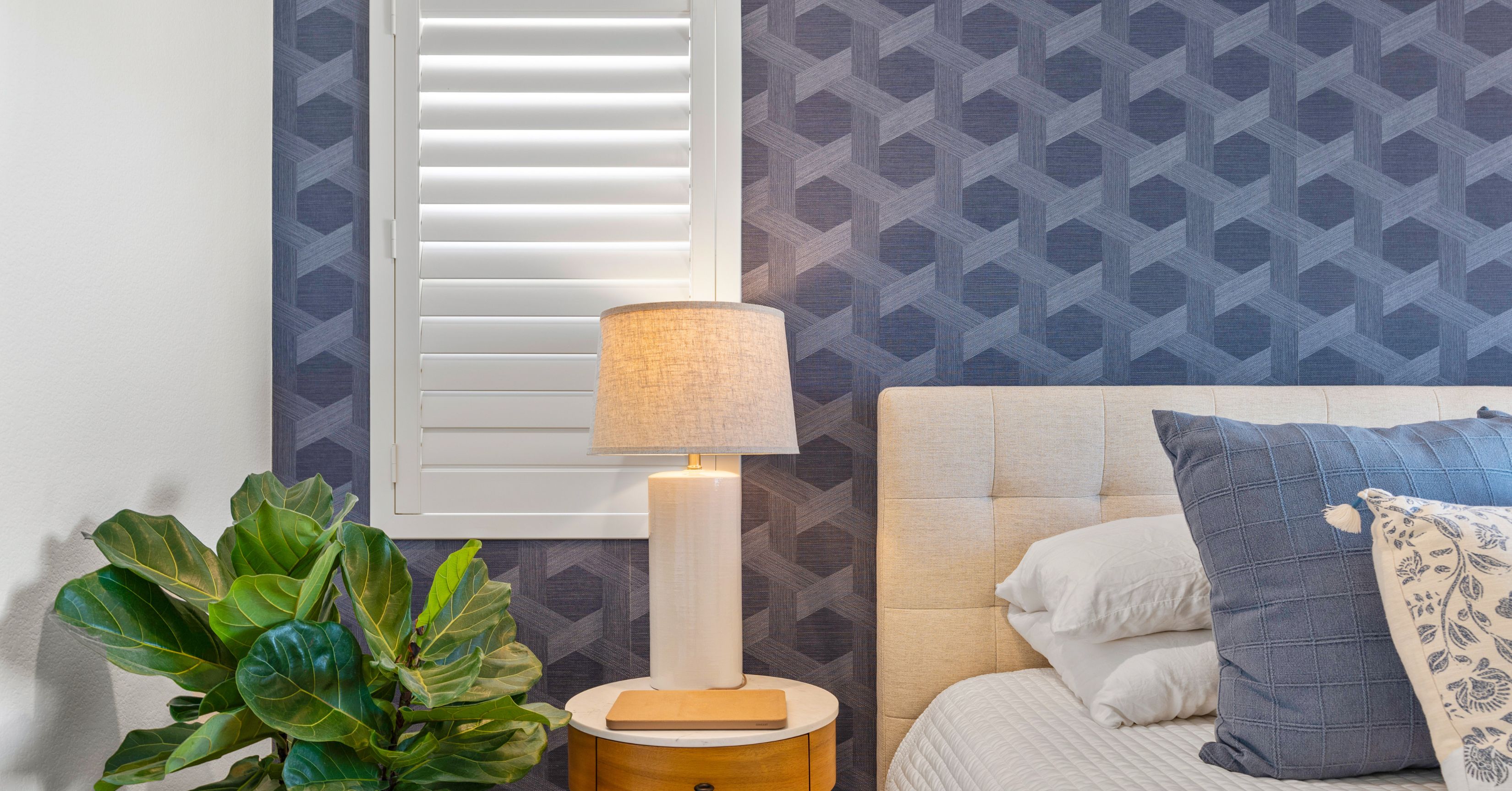 Bedroom with a beige upholstered headboard, navy geometric accent wall, bedside lamp, and leafy green plant.