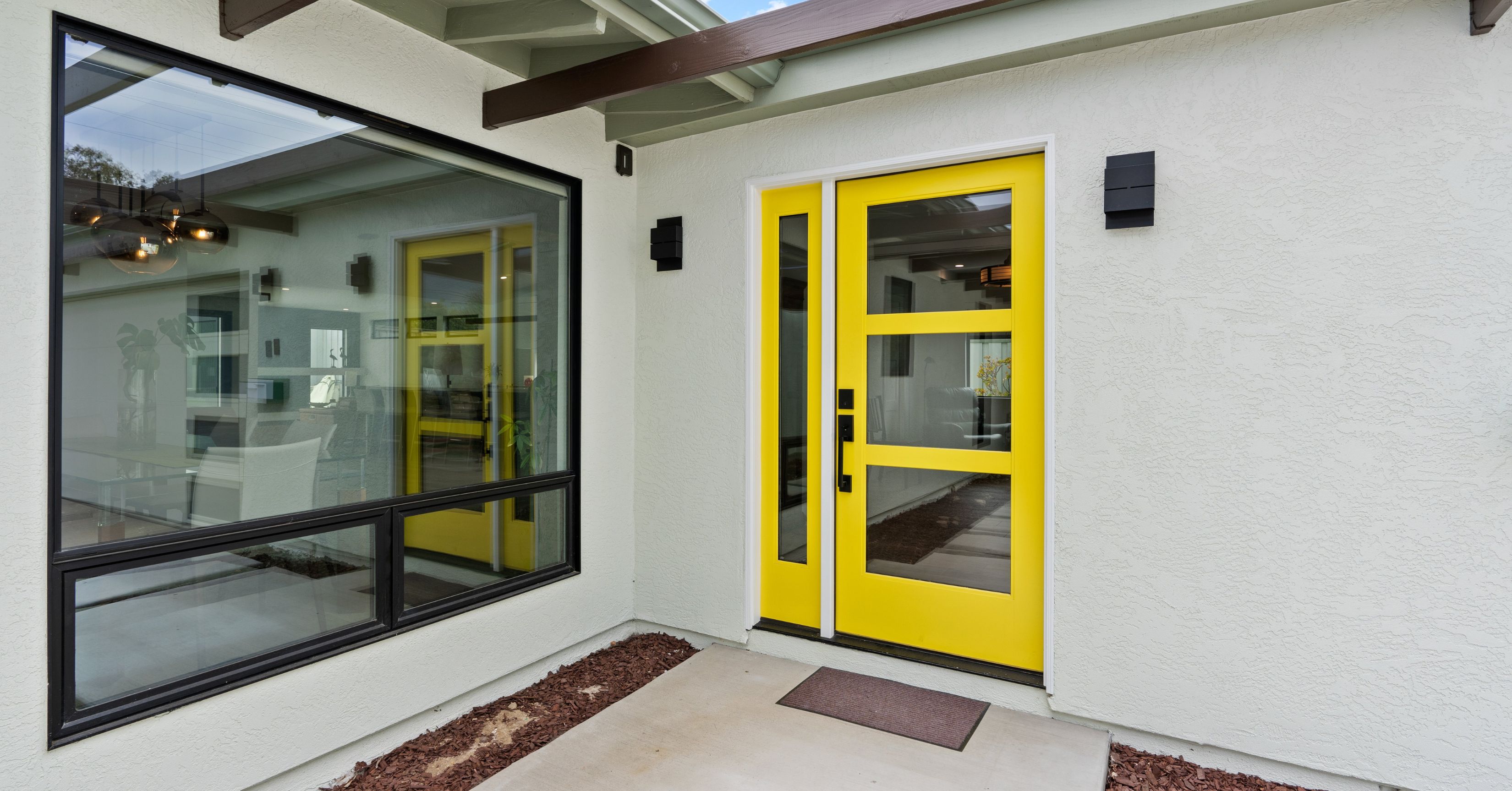 Exterior of an attached ADU addition with a bright yellow glass-paneled entry door, large black-framed window, and white stucco walls.