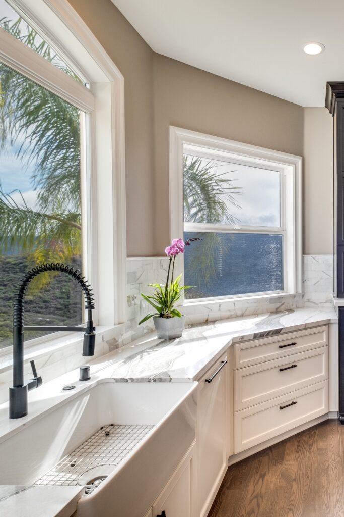 Farmhouse sink beneath large kitchen windows with a black spring faucet, white cabinetry, and veined stone counters.