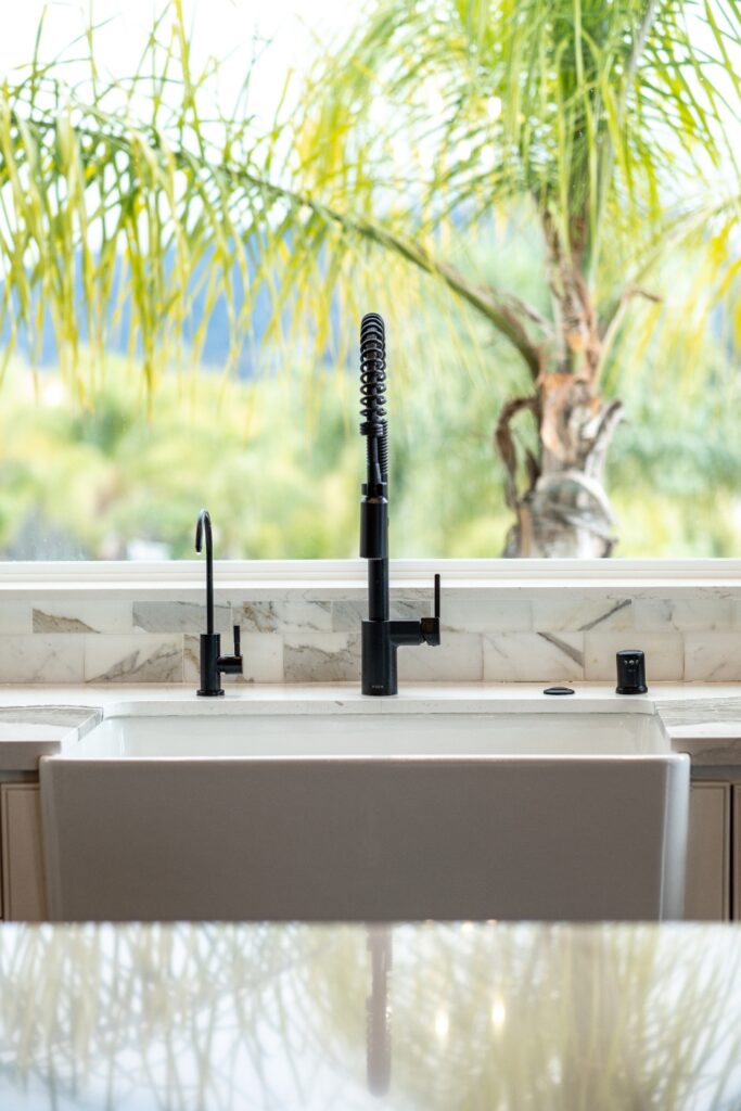 Close-up of a white farmhouse sink with black faucet fixtures and palm trees visible through the kitchen window.