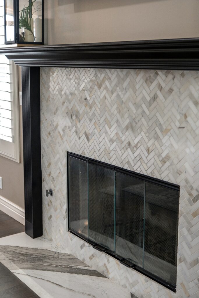 Remodeled fireplace with a black wood surround, herringbone marble tile, glass doors, and veined stone hearth.