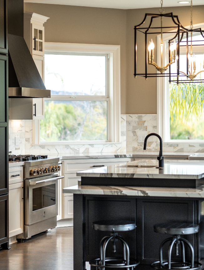 Wide view of an Alpine kitchen remodel featuring a farmhouse sink, black and white cabinetry, veined stone countertops, large windows, and an adjoining dining area.