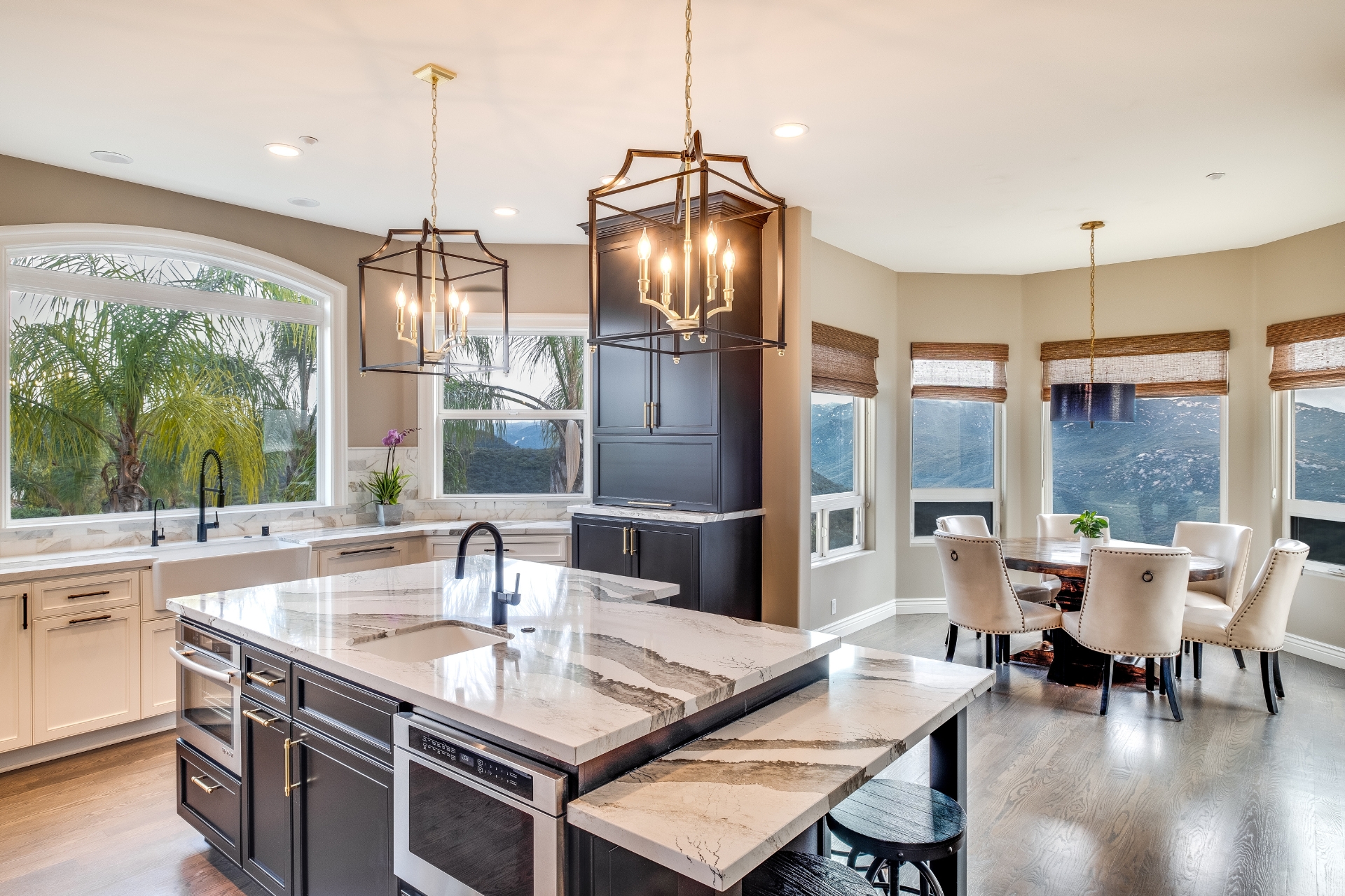 Open Alpine kitchen and dining area with a large stone island, farmhouse sink, black cabinetry, and mountain views through surrounding windows.