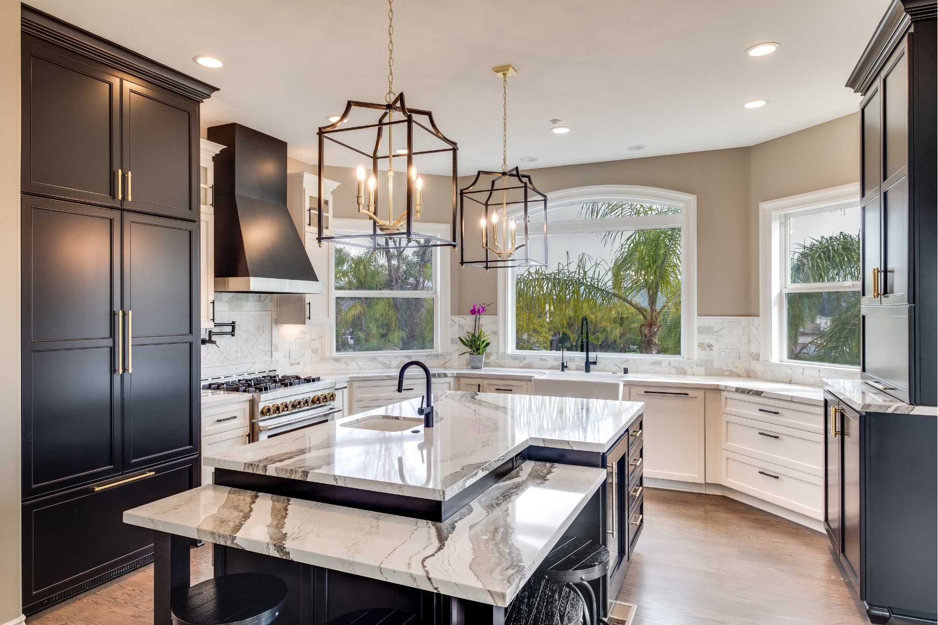 Wide view of an Alpine kitchen with black and white cabinetry, oversized stone island, farmhouse sink, professional range, and large arched window.