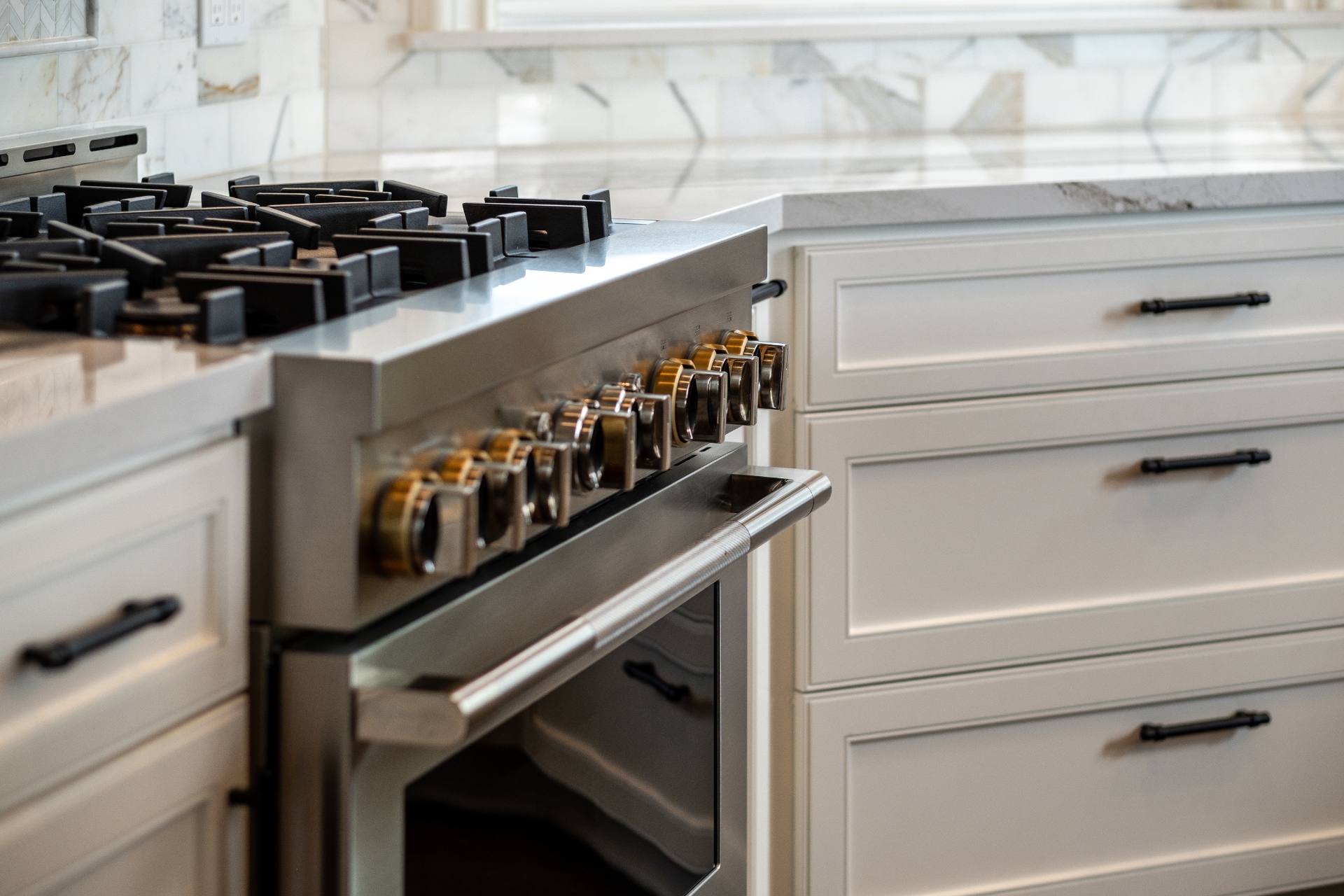 Stainless steel professional gas range surrounded by white shaker cabinets, black hardware, marble backsplash, and stone countertops.