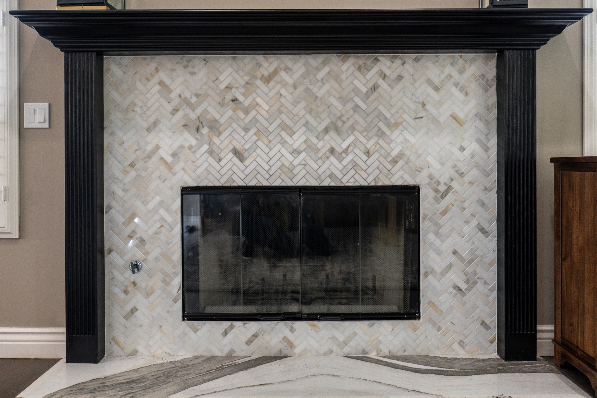 Remodeled fireplace with black wood mantel, herringbone marble tile surround, black glass doors, and veined stone hearth.