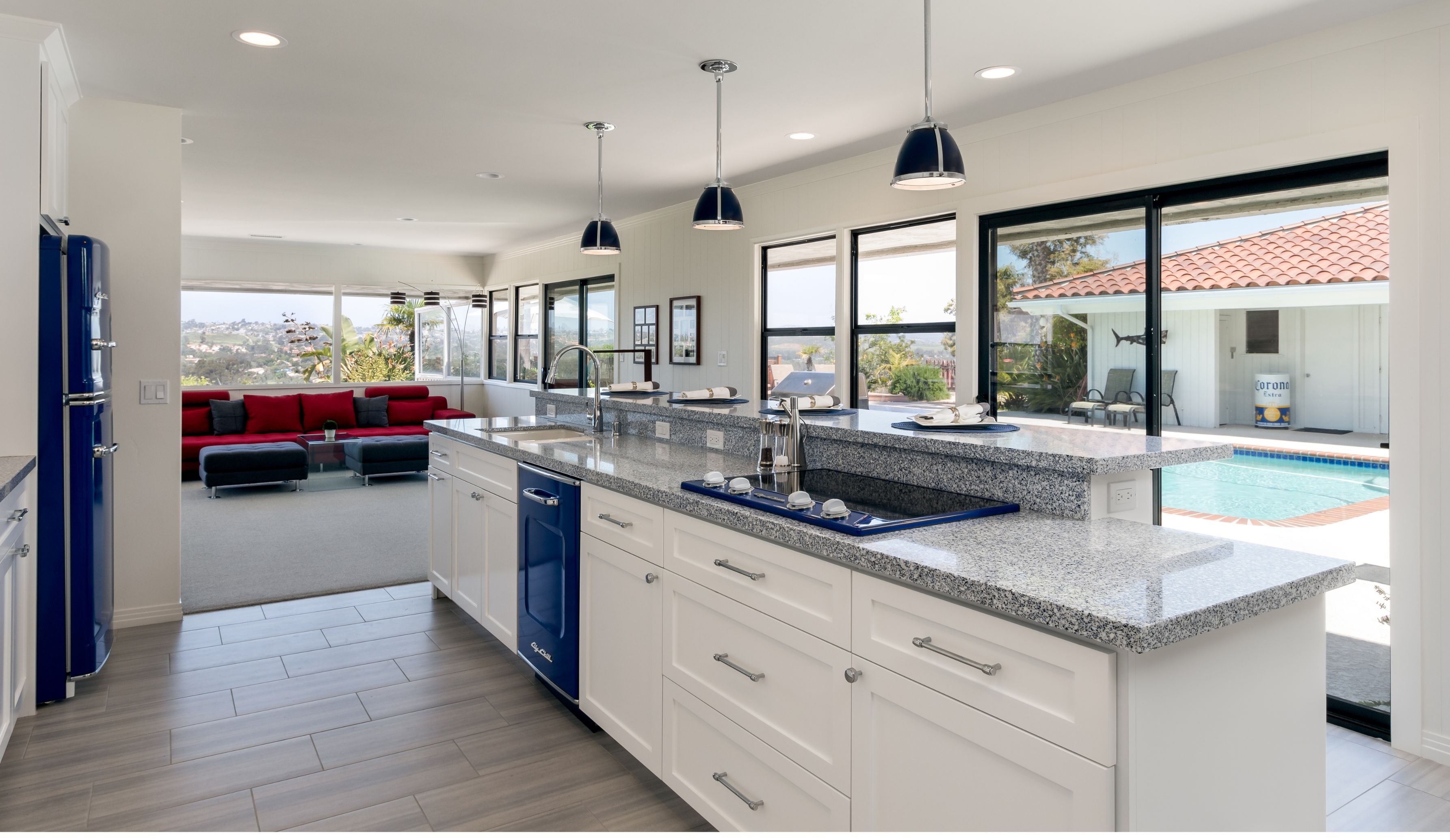 Open Bonita kitchen with a long granite island, sink, cooktop, white cabinetry, blue appliances and pool-facing windows.