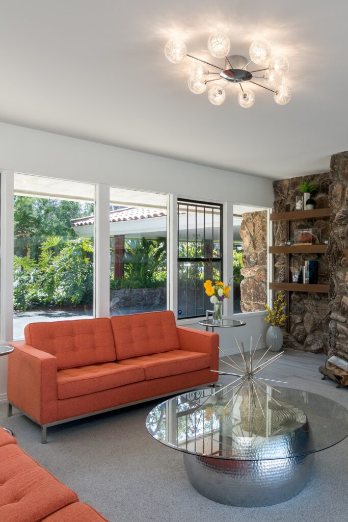 Living room with orange seating, a stone fireplace, floating wood shelves and a round glass coffee table.